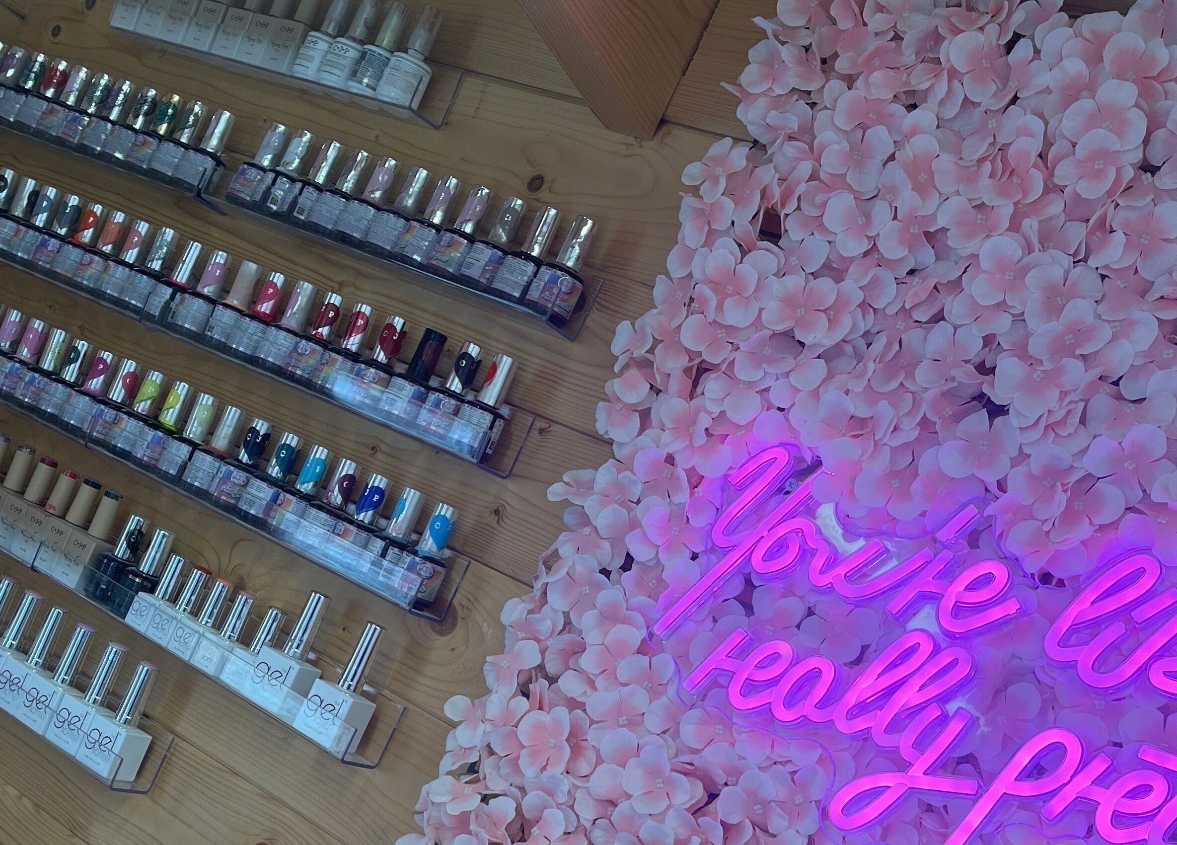 Magpie Nails in Havant, England displays vibrant nail polish rows beside a pink floral wall and neon sign.