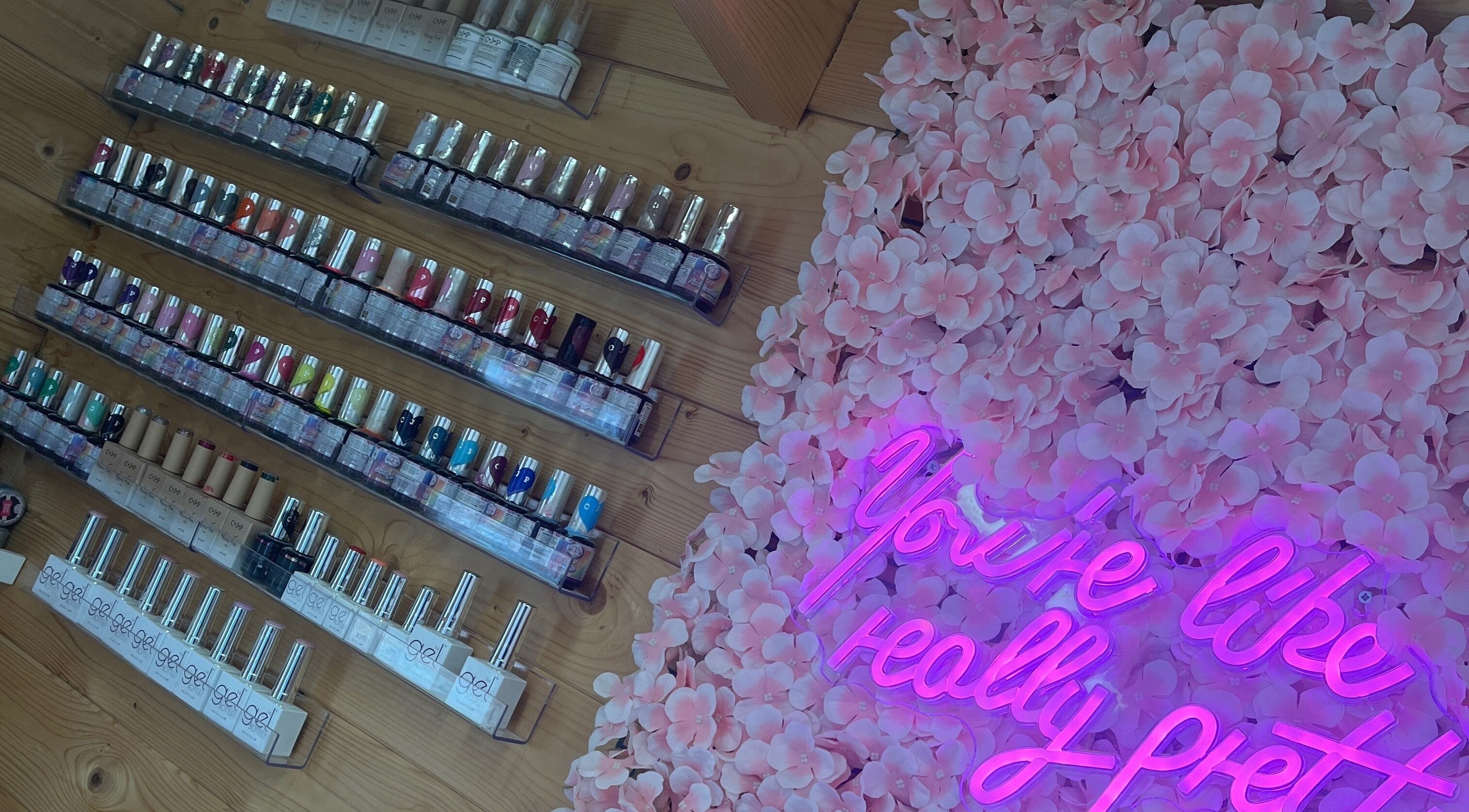 Magpie Nails in Havant, England displays vibrant nail polish rows beside a pink floral wall and neon sign.