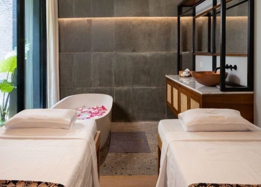 Relaxing treatment room at Ame Spa Bali, Bali, ID with twin massage beds and a petal-filled bathtub.