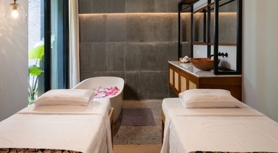 Relaxing treatment room at Ame Spa Bali, Bali, ID with twin massage beds and a petal-filled bathtub.