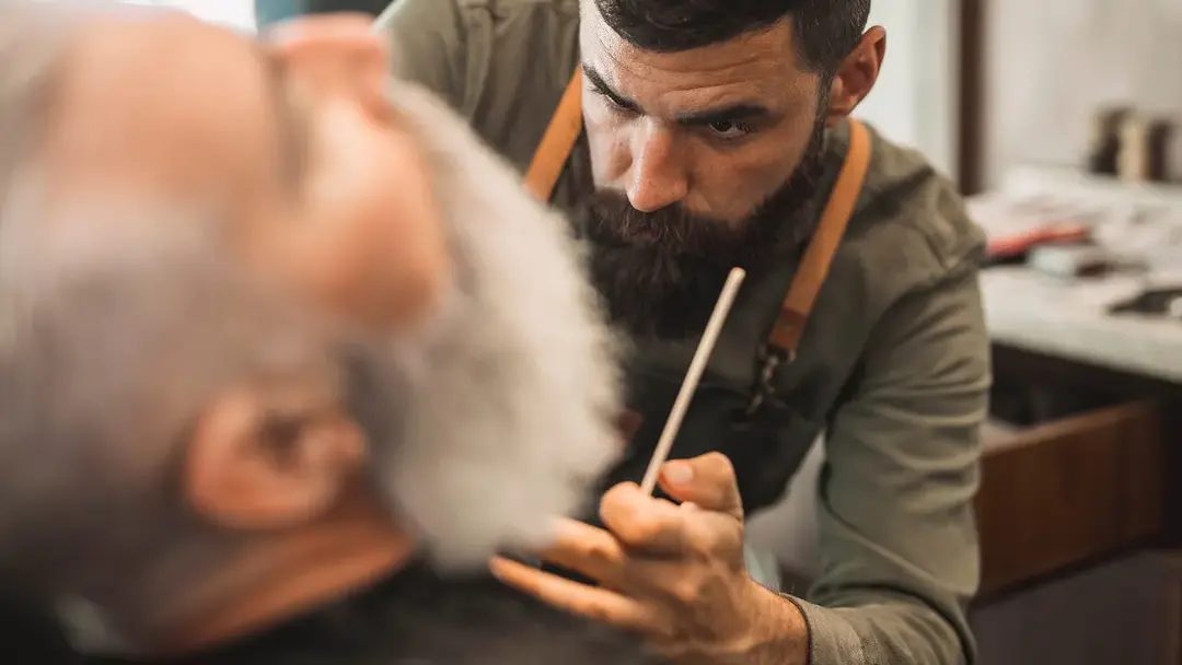 Ein Barbier schneidet mit einer Schere sorgfältig den gepflegten Bart eines sitzenden männlichen Kunden in einem rustikalen Salon.