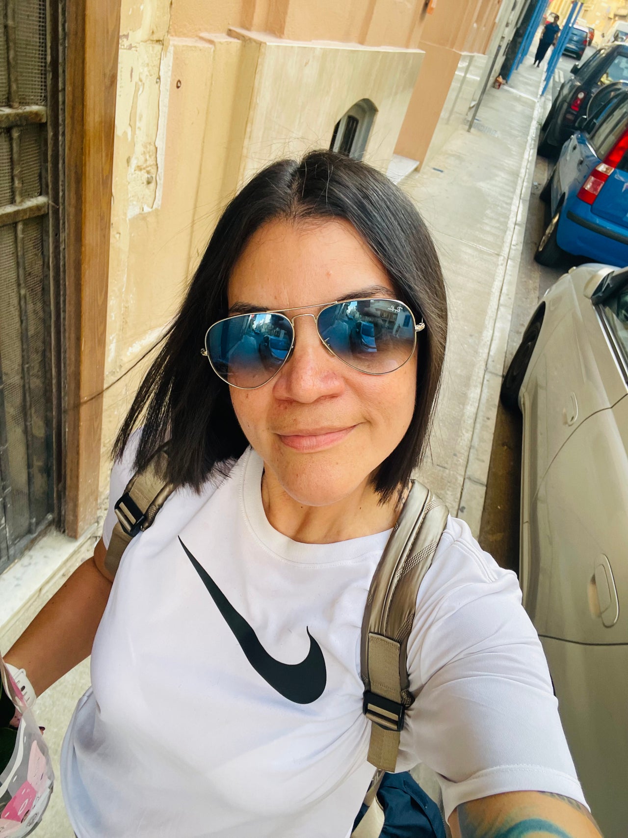 Woman wearing sunglasses and a white Nike shirt stands on a street in front of Arcadiosbarbershop in Tas-Sliema, Malta.